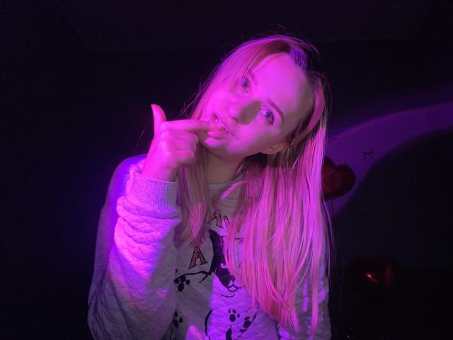 FlyFreedom with pink and purple neon lighting, pointing finger at slightly open mouth, long blonde hair, wearing white hoodie, dark background, red apple partially visible.