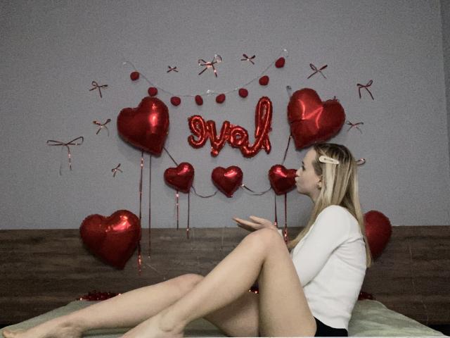 FlyFreedom with long blonde hair, white top, and black shorts, sitting on a bed, legs up, surrounded by red heart balloons and "val" balloon, looking right.
