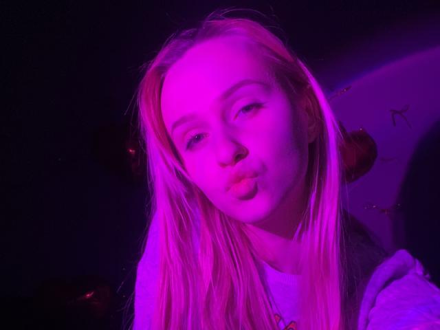 FlyFreedom has pink-tinged blonde hair, pouty lips, and a slightly tilted head, wearing a white shirt, under purple and pink lighting, with a subtle nose ring.