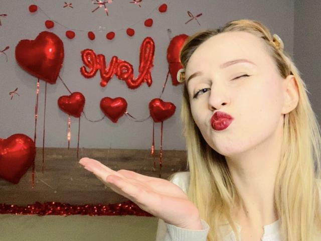 FlyFreedom with pouty red lips, winking, and blowing kiss towards camera, hand up, blonde hair, white shirt, red heart balloons and "snaf" balloon in background.