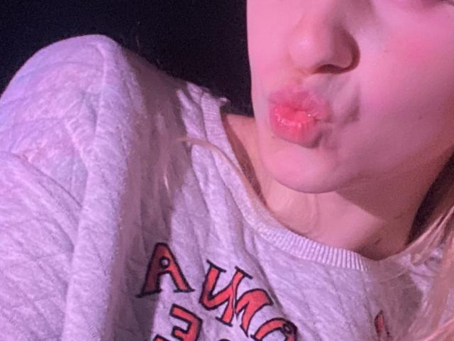 FlyFreedom's pink pout is puckering up with shiny lips, wearing a grey "AWC" sweatshirt. Blonde hair is partially visible. Soft pink lighting highlights her face.