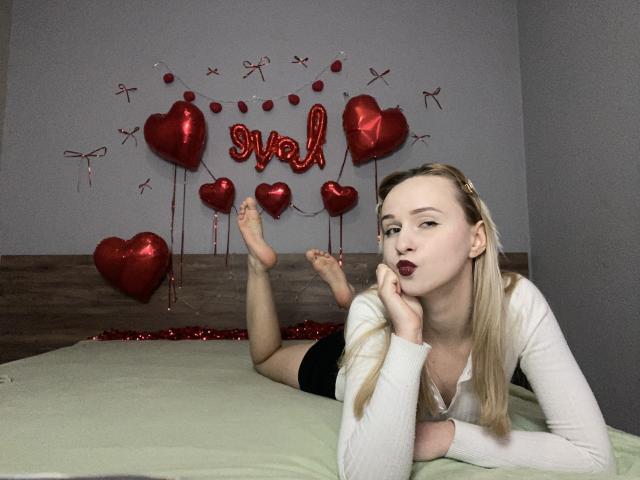 FlyFreedom, blonde, lying on bed, white long-sleeve shirt, black shorts, feet up, looking at camera, red heart balloons, wooden headboard, gray wall, pouty dark lipstick, relaxed.