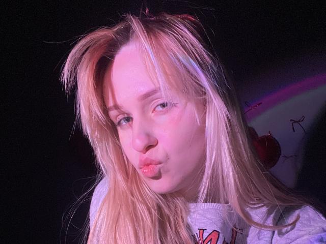 FlyFreedom has blonde hair, pinkish skin, and pouty pink lips, wearing a gray hoodie, with a slight pinkish-purple glow on her face, looking down with a bored expression.