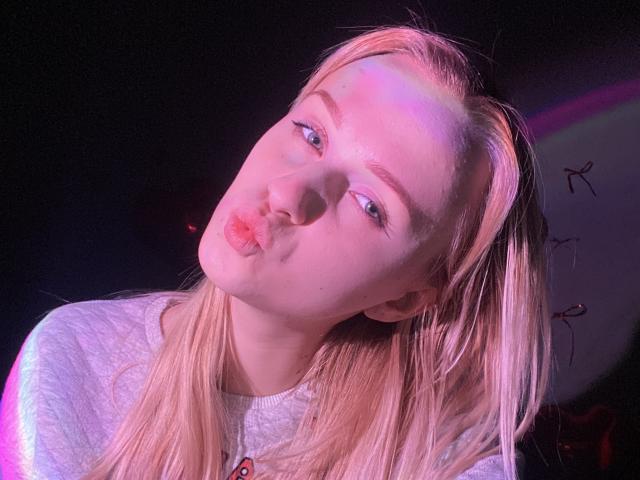FlyFreedom with pouty pink lips, blonde hair, and blue eyes, wearing a white shirt, looking slightly up and to the side, illuminated by purple and pink lighting.