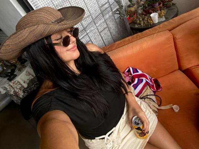 IsabellCollin with long black hair, wearing a brown hat, sunglasses, black off-shoulder top, white shorts, holding a can, and a colorful bag on an orange couch.
