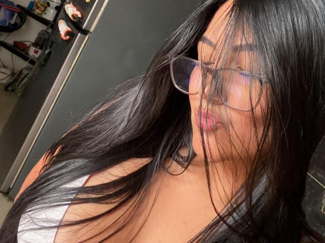 IsabellCollin with long black hair, glasses, and pink lips, her white top showing large boobs, her face partially covered by her hair. She's looking down, slightly blushing.