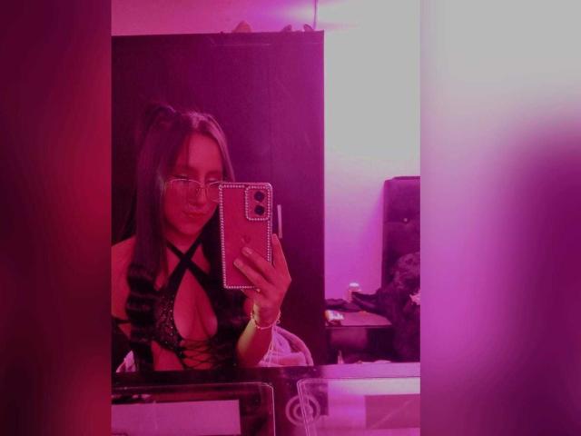 MiaKhalipso with glasses, holding a phone, wearing a black lace-up bra that reveals her big boobs, reflected in a mirror with pink and purple lighting.