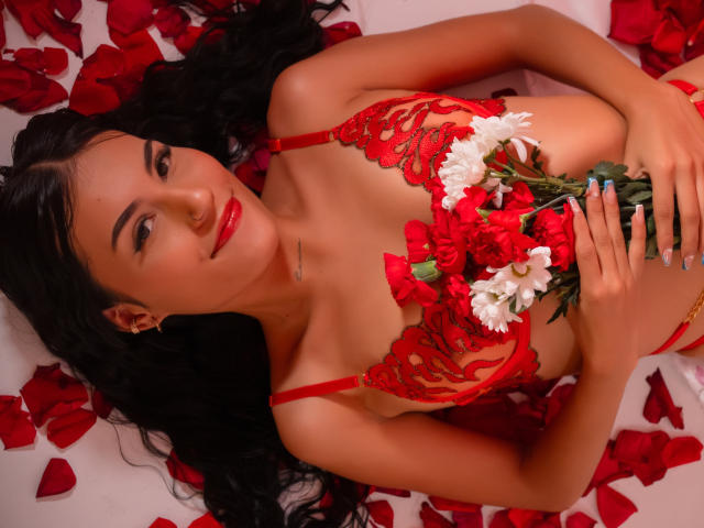BellaTorres with long black hair, red lace lingerie, holding roses and white flowers, lying on red petals, red lipstick, looking up, cute, sexy, medium tits, slim, attractive.