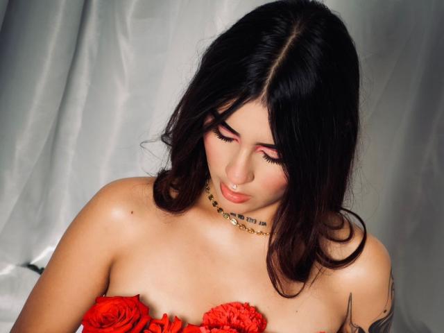 VioletBlazee with dark wavy hair, pink eyelids, nose ring, gold necklace, and red rose-covered tits, looking down, tattoo on right arm.