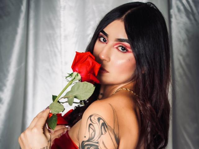 VioletBlazee with long black hair, holding a red rose to her mouth, showcasing a tattoo on her right shoulder, wearing red and gold, looking seductive.