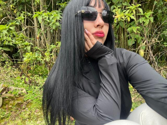 CherrySexxy with long black hair, wearing black sunglasses, a black shirt, and red lipstick, touching her cheek, standing in a green, leafy outdoor setting.