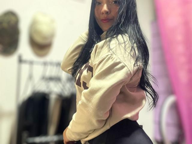 CherrySexxy with long black hair, wearing a beige sweatshirt and black high-waisted pants, looking over her shoulder with a subtle smile. Boobs not visible, ass not visible.