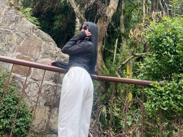 CherrySexxy with long black hair, black top, and white pants, leaning on a rusted metal railing, showing her ass, wearing sunglasses, in a green, rocky, forested area.
