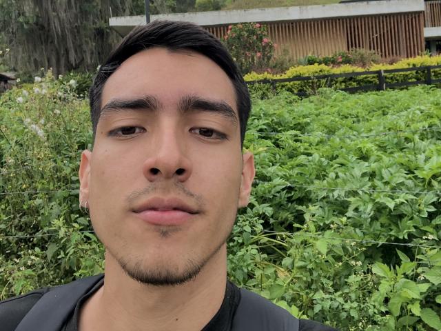 NicholasFinn has short dark hair, a slight mustache, and a neutral expression, wearing a black shirt, with green foliage in the background. His skin is light brown.