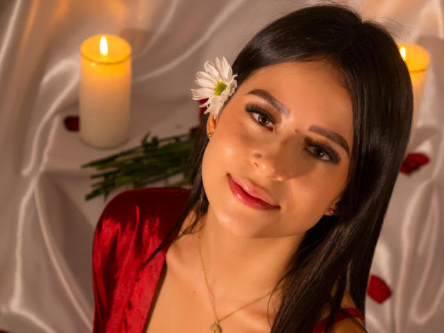 MadissonBlue, con cabello oscuro, vestido rojo, una flor blanca en el cabello, sonriendo, ojos marrones, lápiz labial rojo, iluminación con velas, luz suave, maquillaje sutil, collar, brillo sutil, relajada, sensual, atractiva..
