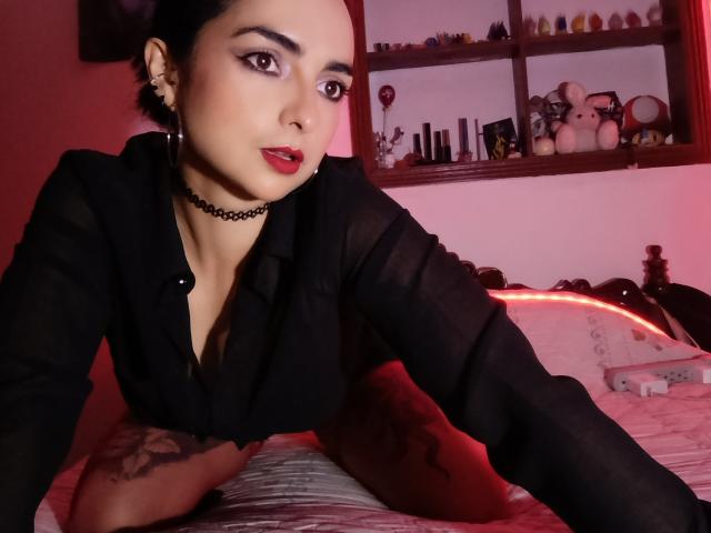 CandyParis with dark hair, red lipstick, black sheer blouse, choker, and tattoo on arm, leaning on bed, red lighting, looking at camera. Boobs slightly visible through blouse.