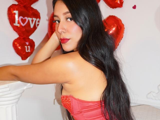 LiahSantorini with long black hair, red lipstick, and red lingerie, showing her ass, leaning on a white column, red heart balloons in the background.