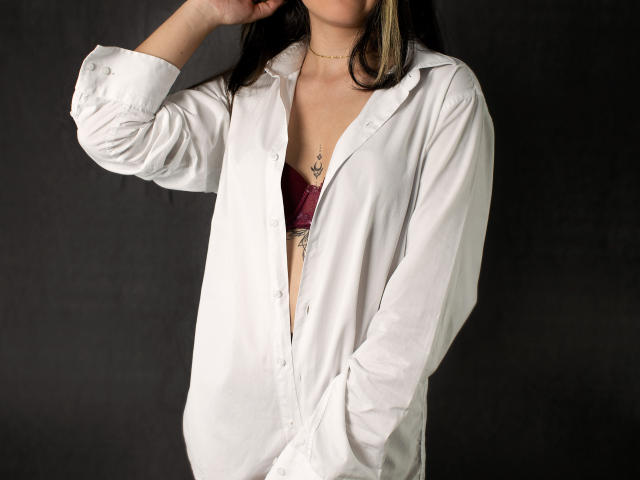 KatieDesirX in a white button-up shirt, hands in pockets, showing a red bra with small tits and a chest tattoo, dark hair, black background.
