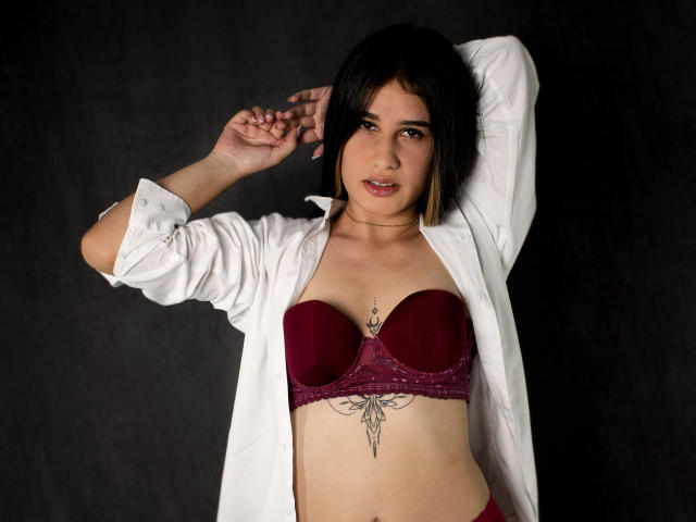 KatieDesirX with black hair, wearing an unbuttoned white shirt, maroon bra, and matching panties, exposing a tattoo below her tits, arms raised, dark background.