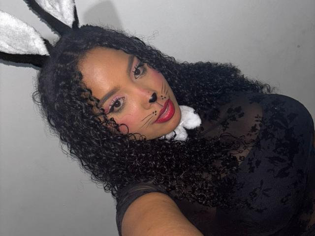 CarolinEmml with curly black hair, black sheer top, white bunny nose, black bunny ears, pink eyeshadow, red lipstick, and white furry collar, looking at camera, gray background.