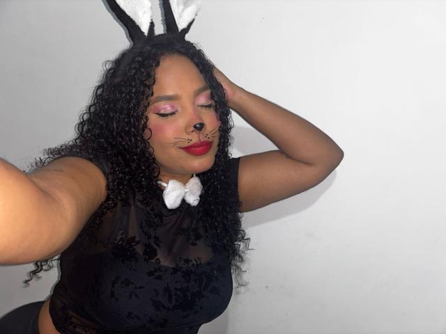 CarolinEmml with curly black hair, pink eyeshadow, red lipstick, black lace top, white bunny ears, and white bow, selfie showing her face and arm raised. Boobs, pussy not visible.
