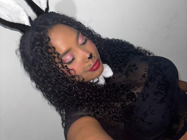 CarolinEmml with curly black hair, pink eye makeup, black lace top, white collar, black bunny ears, and red lipstick, eyes closed, showing off her large, round boobs.