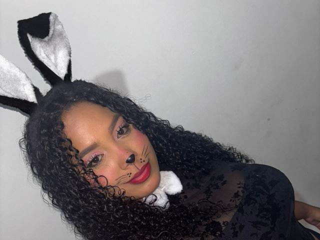 CarolinEmml with curly black hair, wearing black sheer top, white collar, and black/white bunny ears, has pink makeup, whiskers, and red lipstick, looking at camera.