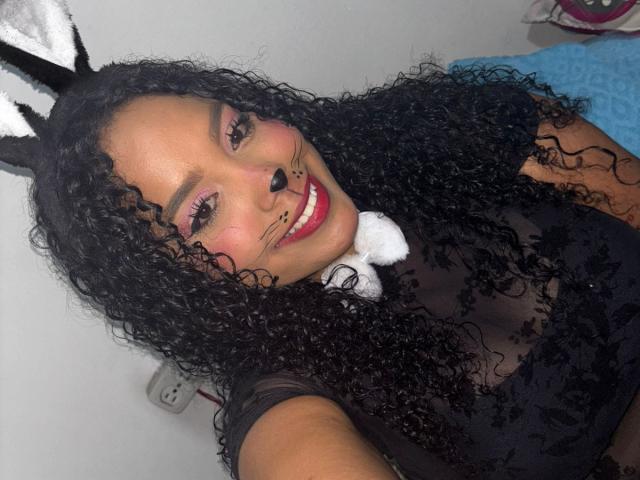CarolinEmml with curly black hair, wearing black lace top, bunny ears, and black nose, smiling with red lipstick, pink eye makeup, and white fluffy collar. Boobs visible through lace.