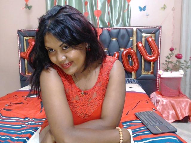 FelyLove with dark brown skin, black wavy hair, red lace dress, smiling at camera, sitting on red and blue striped bed, wearing red earrings and beige bracelet.