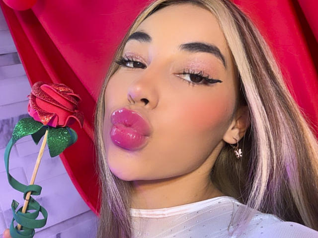 MissVallen with light brown, straight hair, full pink glossy lips, and dark winged eyeliner, holding a glittery red rose on a green stick. She's wearing a white sheer top with small silver dots. A small pink star earring is visible. Her skin tone is light brown. The red curtain background is slightly out of focus. The image is a close-up portrait.