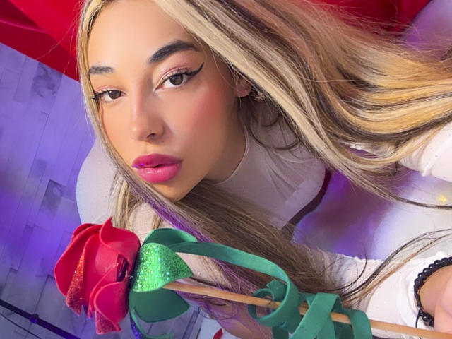Close-up of MissVallen with long, straight blonde hair, pink lipstick, and winged eyeliner. She holds a rose with a green glittery stem and green ribbon. Her white shirt contrasts with the red background. Her expression is neutral, slightly curious. The composition centers on her face and the rose, with the rose's vibrant colors drawing attention. The image has a modern, digital look.