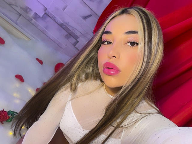 MissVallen with long, straight, blonde-highlighted hair and pink lipstick. She wears a white, sheer, long-sleeve top with small polka dots. Her eyebrows are dark and well-defined. Rose petals are scattered on the floor. Red curtains and a gray, textured wall are in the background. The lighting is soft, with a slight purple hue. Her makeup is bold, emphasizing her eyes and lips. She's looking directly at the camera.