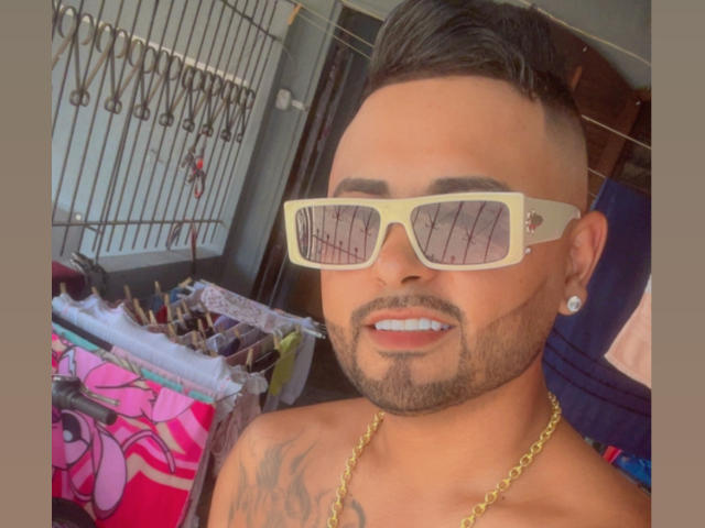 Morenoses with tan skin, black undercut, and beard, wearing yellow sunglasses, gold chain, and ear piercing, showing off a tattoo on his right shoulder. Boobs not visible.