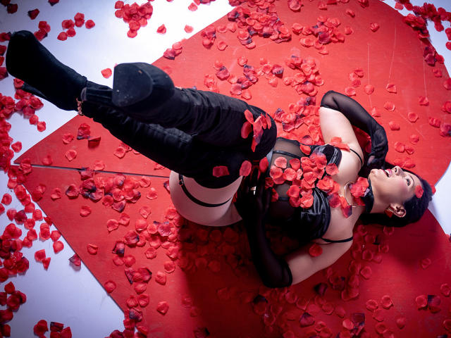 AdaMclain lying on red mat, surrounded by rose petals, wearing black latex outfit, gloves, and boots, with rose petals covering her tits and pussy, looking up.