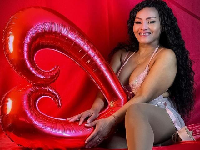 AmbarBlack with long black curly hair, smiling, wearing pink lace lingerie and sheer pantyhose, holding a shiny red "3" balloon, sitting on red fabric. Boobs visible, ass partially seen.
