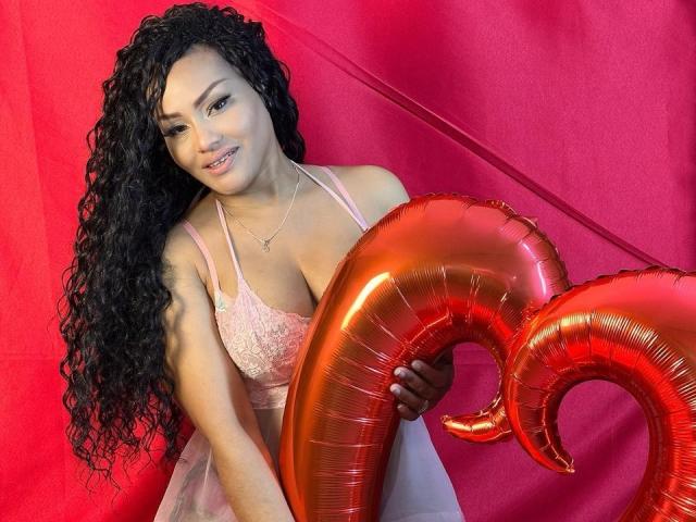 AmbarBlack has long curly black hair, wearing a pink lace bralette, holding a shiny red balloon shaped like a curled horn. She's smiling with light brown eyes and a pink lip gloss. The red background contrasts with her pink outfit and the balloon. AmbarBlack's nails are painted white. The image has a playful, celebratory vibe with a focus on bright colors and shiny textures.