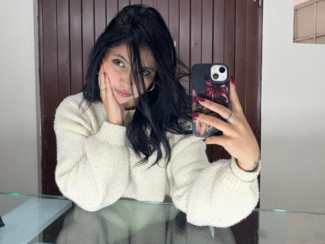 NyraVanel with black hair, red nails, and a white sweater, holding a phone with a pink flower case, smiling at a mirror, touching her cheek.