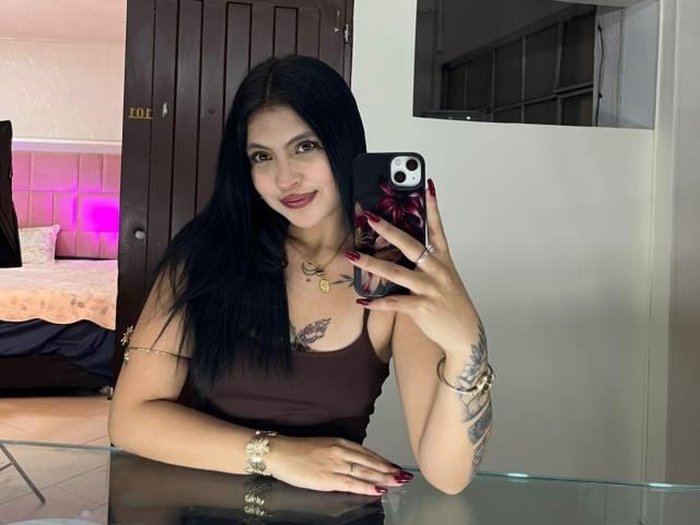 NyraVanel, con cabello largo y negro, un top marrón, uñas rojas, tatuajes en los brazos, un collar dorado y una funda para el teléfono, tomando una selfie frente a un espejo en una habitación con una cama..