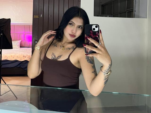 NyraVanel with black hair, brown tank top, tattoos on chest and arm, holding phone, mirror selfie, showing tits, red nails, gold bracelet, in room with bed and door.