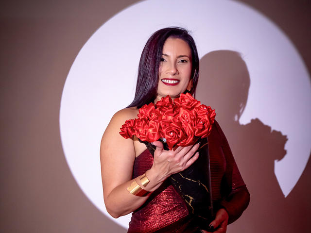 AdaMclain with red roses, holding bouquet, smiling, wearing red dress, gold bracelet, dark hair, shadow on wall, looking at camera, showing tits.