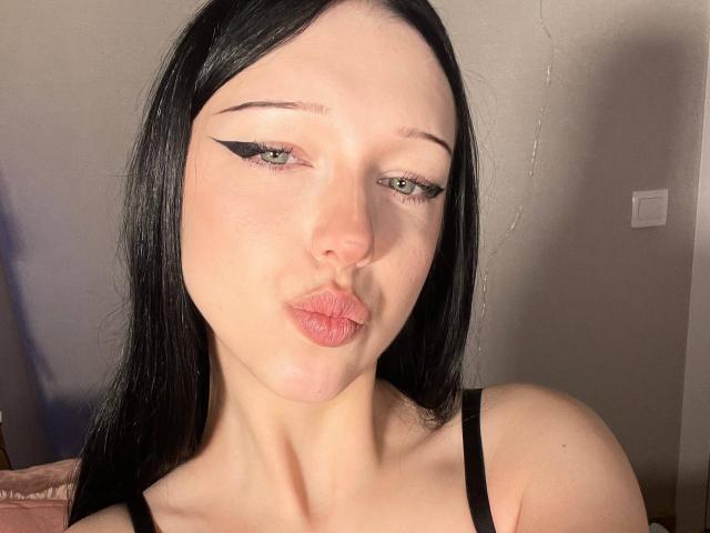 AmaiRin with pale skin, black straight hair, and sharp black eyeliner, pouting her pinkish lips. She's wearing a black spaghetti strap top, looking seductive.
