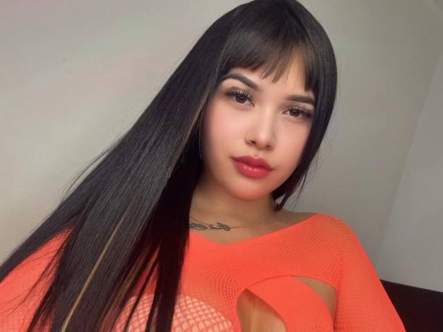 LunaAndKlaus with long black hair, bangs, and red fishnet top, showing cut-out sections, revealing her tits, has a tattoo on her chest, and red lipstick.