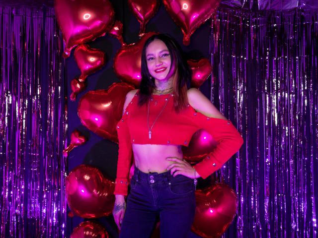 LiiaSmith stands in front of a backdrop of shiny purple streamers and red heart-shaped balloons. She wears a red, long-sleeve crop top with sparkling details, exposing her midriff, and high-waisted black jeans. LiiaSmith has straight, dark brown hair, light skin, and is smiling with red lipstick. Her right hand is on her hip, and she wears a necklace with a small cross. The lighting is purple, highlighting the shiny textures.