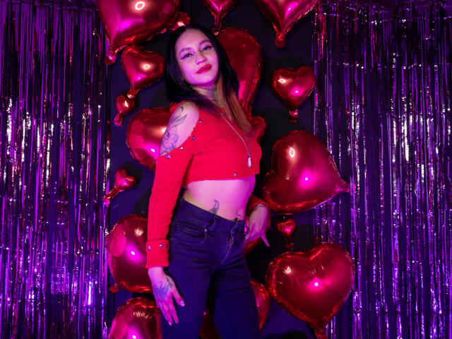 LiiaSmith with red heart balloons, wearing a red crop top, black jeans, and showing tattoos, has red lipstick and long brown hair, standing in front of shiny purple streamers.