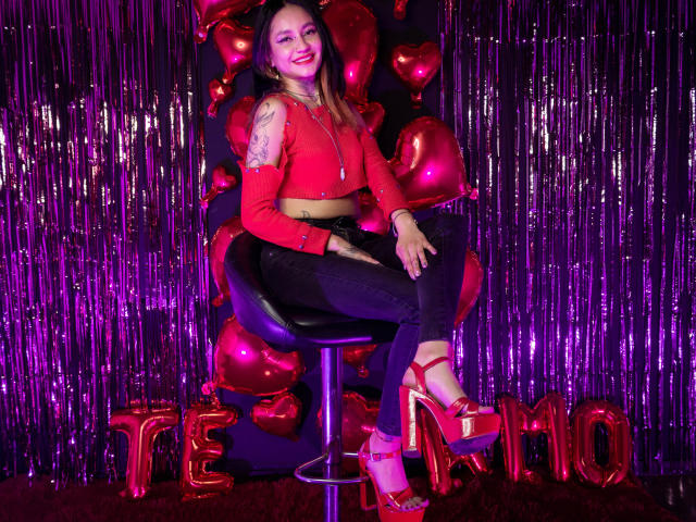 LiiaSmith with pink lipstick, red crop top, black jeans, and red platform heels, sitting on a stool, smiling, showing her tattooed arm, surrounded by red heart balloons.