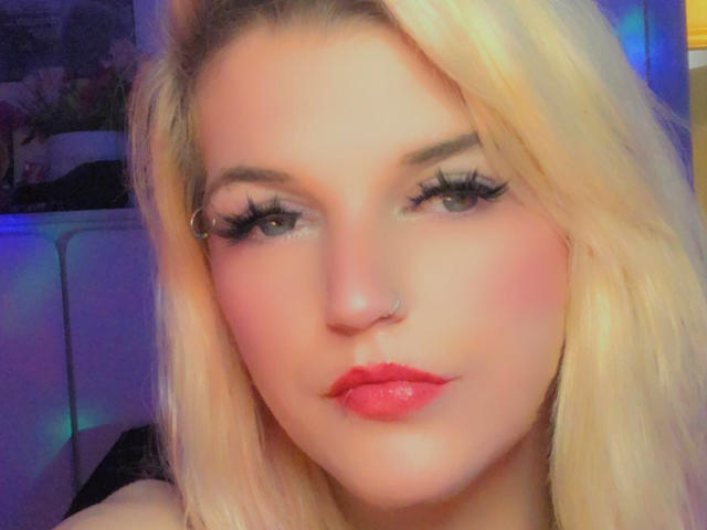 StoneySparx69 with blonde hair, nose ring, and pink lipstick, sporting long eyelashes, looking at the camera with a slight pout, in a well-lit, colorful setting.