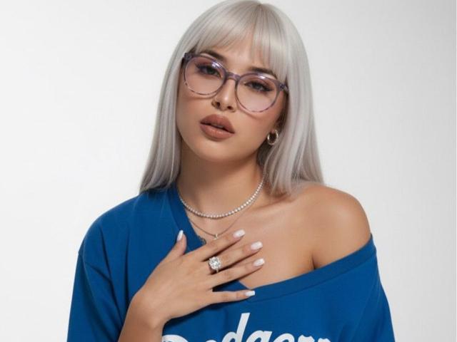 LunaAndKlaus with silver hair, big glasses, and blue shirt, off-shoulder, showing tits, hand on chest, ring, necklace, white nails, and gold earrings.