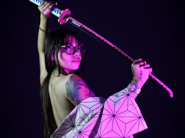 PrincessMiley holds a katana with a white hilt and black blade, positioned diagonally across her body. She has long black hair with bangs, glasses, and a tattoo of a woman's face on her left arm. She's topless, with a geometric-patterned cloth covering her lower body. Her right arm is raised, and her left arm is slightly bent. The lighting is purple, highlighting her tattoo and cloth. Her expression is confident.