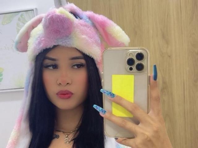 LunaAndKlaus with dark hair, pink lipstick, and colorful fluffy bunny hat, holding a phone with yellow grip, blue nails, in a mirror selfie.