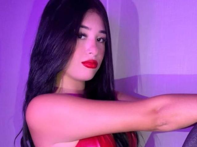 LunaAndKlaus with long black hair, red lipstick, and red top, arms crossed, looking at camera, purple light, sexy, confident, full lips, dark eyes, subtle makeup.